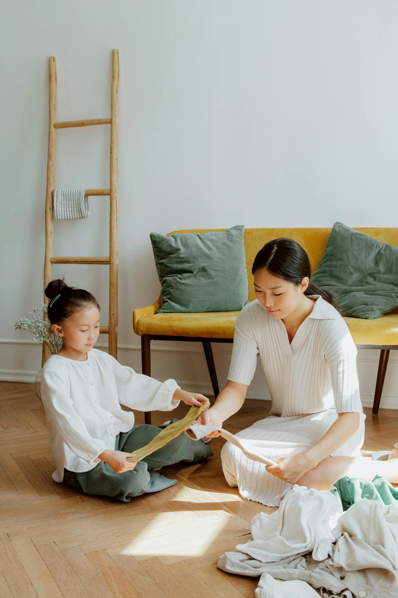 A mother and daughter smiling while folding laundry indoors on a sunny day.